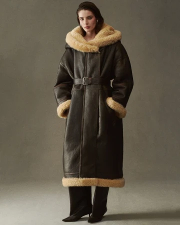 Model wearing a long dark brown shearling coat with plush cream trim and a belted waist against a neutral grey studio backdrop.