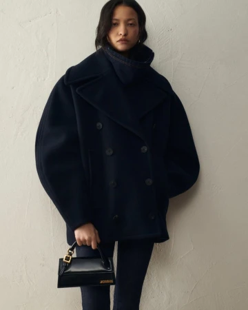 Model posing in an oversized navy pea coat and holding a black structured leather handbag against a textured off-white wall.