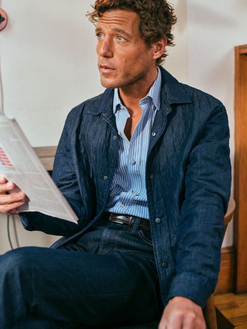 Man in denim jacket and jeans sitting indoors, reading newspaper in a casual pose, with soft lighting and a classic style.