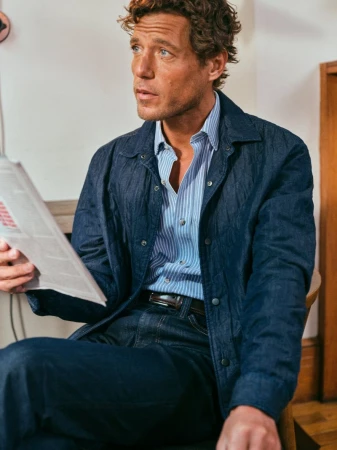 Man in denim jacket and jeans sitting indoors, reading newspaper in a casual pose, with soft lighting and a classic style.