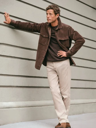 A stylish adult man leans against a grey wall, wearing a brown corduroy jacket, dark brown t-shirt, beige pants, and brown suede shoes in an outdoor setting.