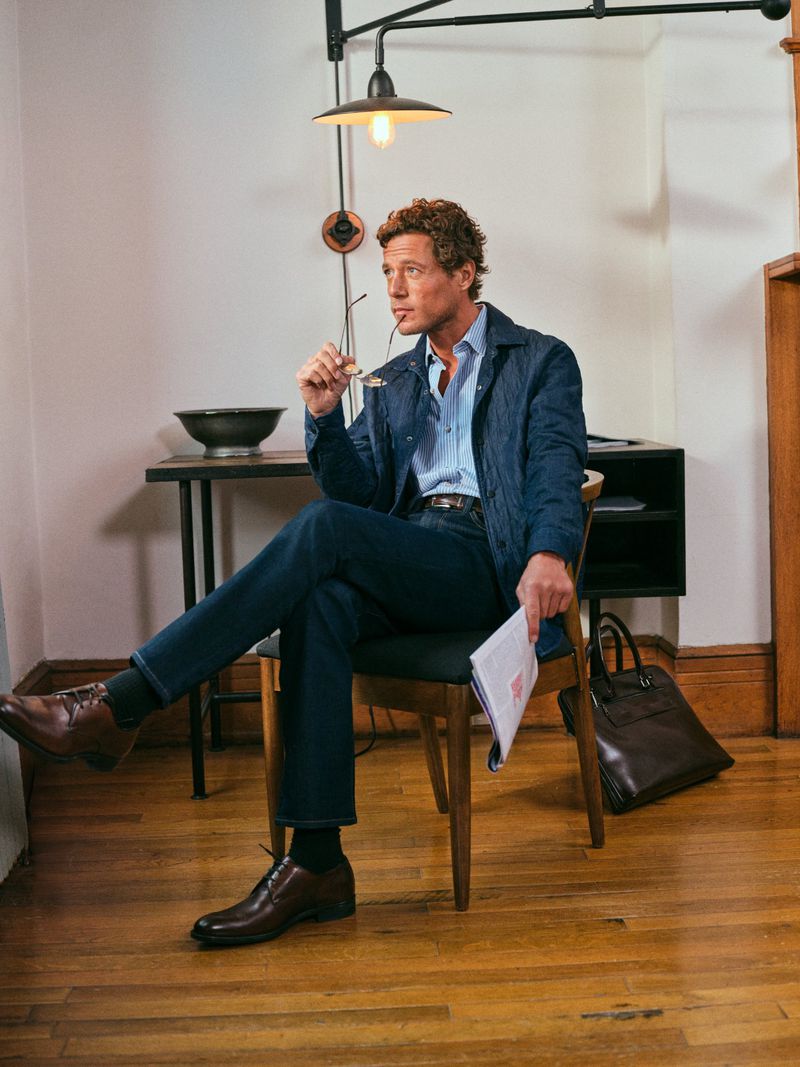 Man in blue jacket and jeans sitting on chair indoors, holding glasses and paper, wearing brown shoes. Interior setting.