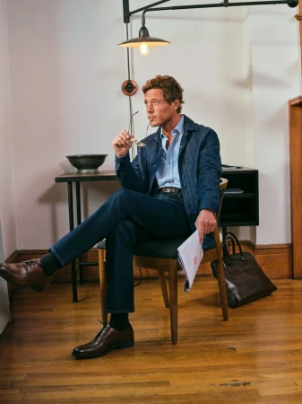 Man in blue jacket and jeans sitting on chair indoors, holding glasses and paper, wearing brown shoes. Interior setting.