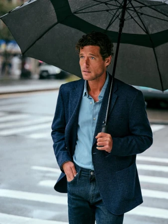 Man in blazer with an umbrella standing on city street crosswalk on a rainy day, wearing dark jeans and button down shirt.