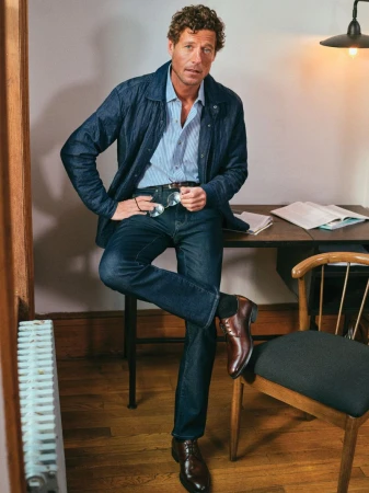 A mature adult male dressed in a denim jacket, jeans, and leather shoes is posed indoors in a casual fashion shot with books and a chair.