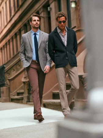 Two adult men walking on a city street, one wearing a blazer and tie, the other sunglasses and a jacket, with buildings in the background.