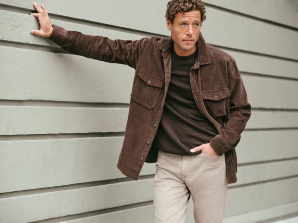A curly-haired adult male poses leaning against a gray horizontal-panel wall, wearing a brown corduroy shirt and tan pants.