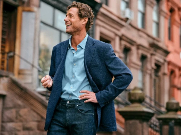 Man in a blue blazer and jeans stands smiling on a city street near brownstones, creating a lifestyle fashion portrait.