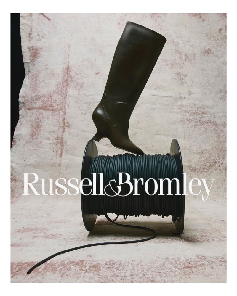 A tall dark leather boot is staged upright atop a spool of dark cord, with the Russell & Bromley logo in the foreground.