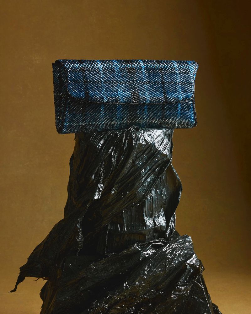 Still life shot of a blue plaid clutch bag on a crinkled black plastic stand, set against a solid gold background. Fashion still life.