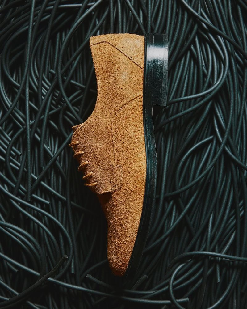 Tan suede dress shoe is presented on a nest of tangled black cables, creating a textured and visually contrasting product shot.