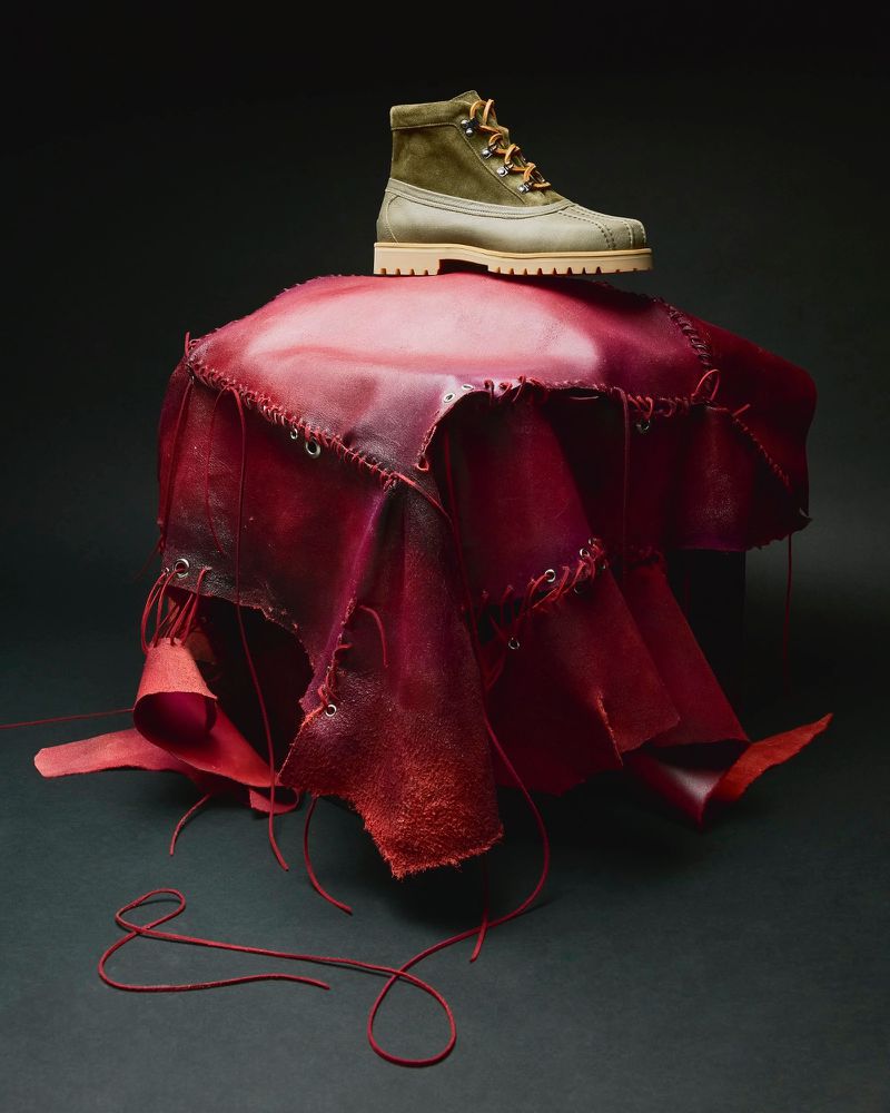 High-angle product shot of a light-brown and olive green boot placed atop a red leather-draped object against a black background.