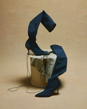 Elevated fashion shot featuring thigh-high, pointed-toe black boots artfully arranged on a fabric-covered platform against a tan backdrop.