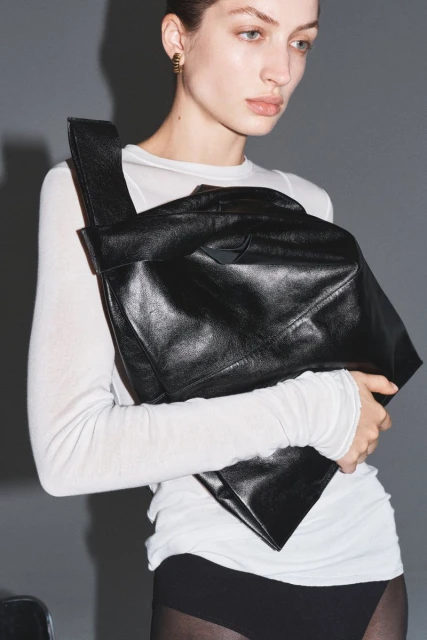 A model presents a large black leather hobo bag, accessorizing a sheer white long-sleeve top and black bottoms, in a studio setting.