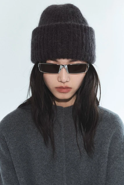 Close-up of an adult woman wearing sunglasses, a gray sweater, and a dark gray knit hat against a bright white background indoors.