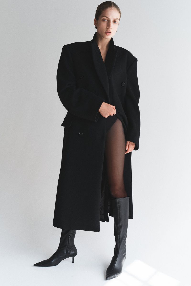 A woman with fair skin poses indoors in a long, black, double-breasted wool coat and black boots against a neutral backdrop.