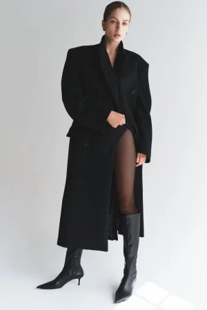 A woman with fair skin poses indoors in a long, black, double-breasted wool coat and black boots against a neutral backdrop.