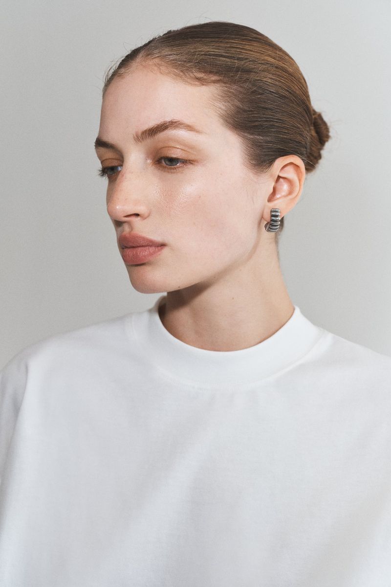 Close-up portrait of an adult woman with neutral expression wearing a white top and hoop earrings against a neutral backdrop.