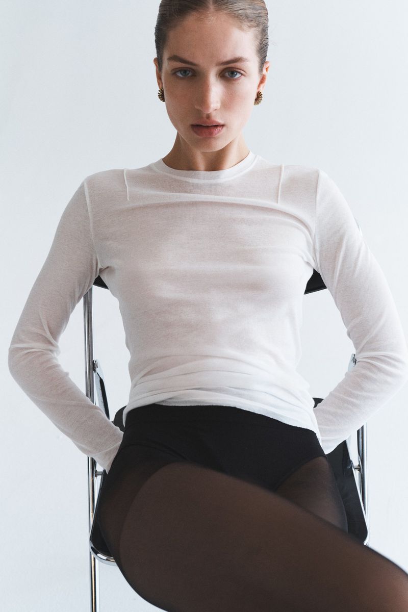 Model Sydney Stabreit seated on a chair wearing a sheer white long-sleeve top and black tights against a plain white studio backdrop.
