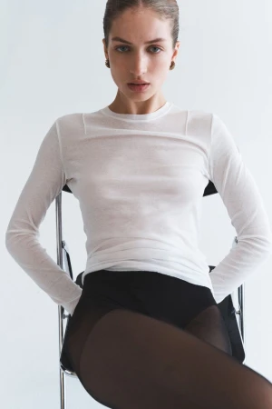 Model Sydney Stabreit seated on a chair wearing a sheer white long-sleeve top and black tights against a plain white studio backdrop.