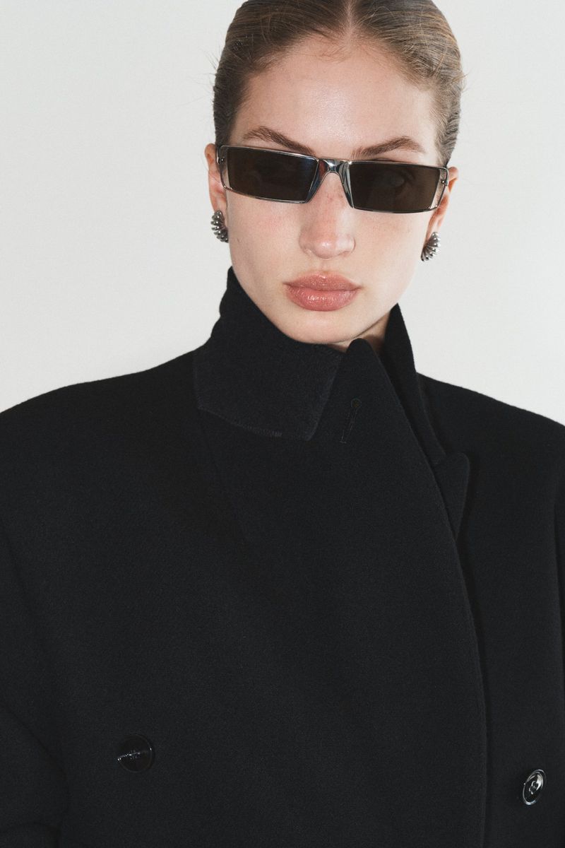 Model wearing dark sunglasses and a black coat against a plain white background
