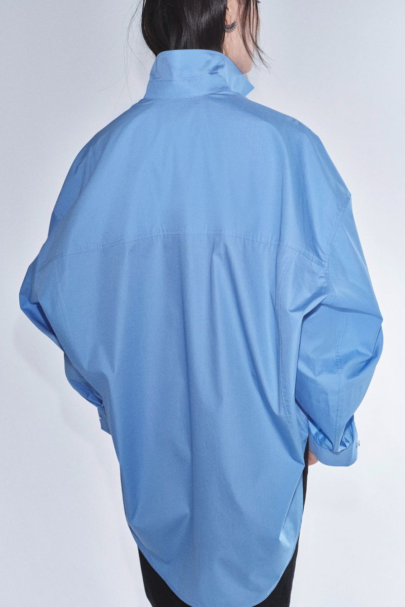 Back view of an unrecognizable fashion model in a blue oversized shirt, standing against a white studio backdrop.