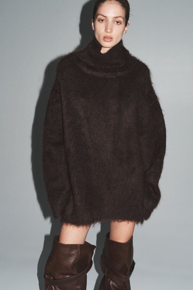 Woman in studio wearing oversized brown sweater dress and high brown boots against a light-blue background in a fashion editorial shot.