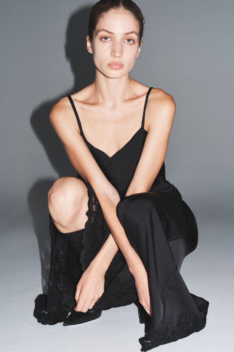 Elegant adult woman in a black slip dress posing in a photo studio. The dress has lace trim. She is sitting on the floor.