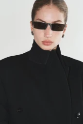 Model wearing dark sunglasses and a black coat against a plain white background