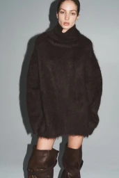 Model wearing an oversized dark fuzzy turtleneck sweater dress and slouchy knee-high leather boots against a grey studio backdrop.