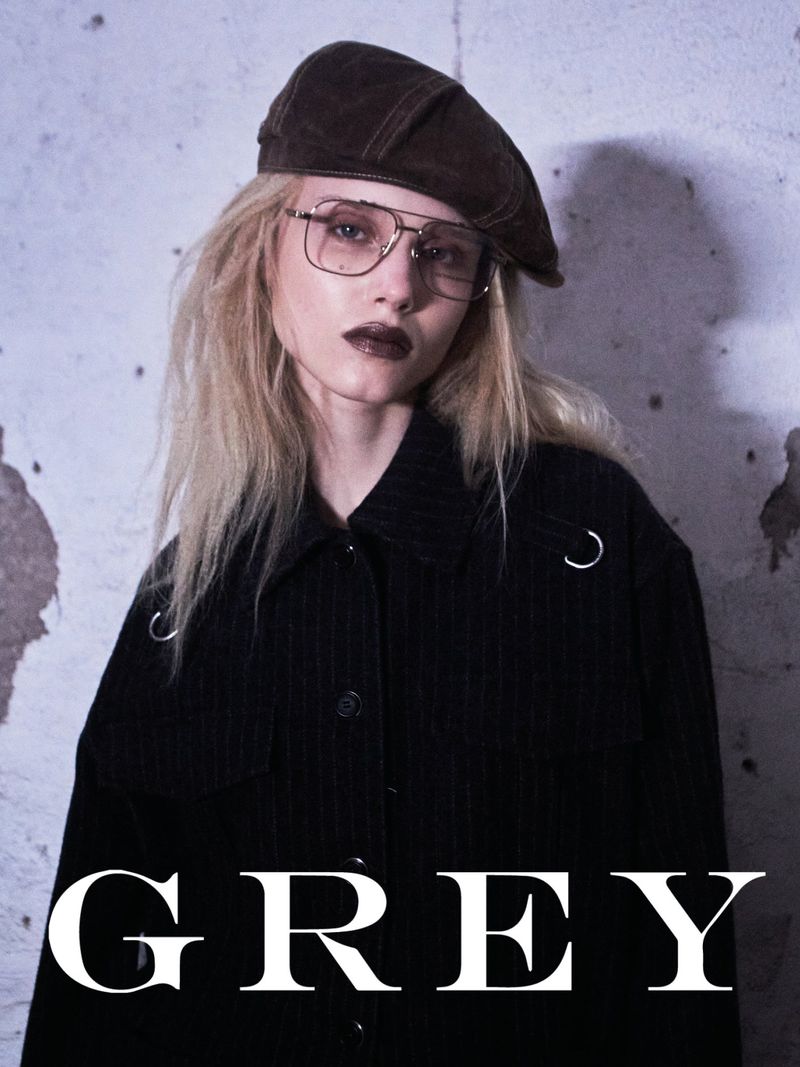 Close-up of a blonde adult female posing indoors. She wears a beret, glasses, dark lipstick, and a pinstripe jacket against a concrete wall.