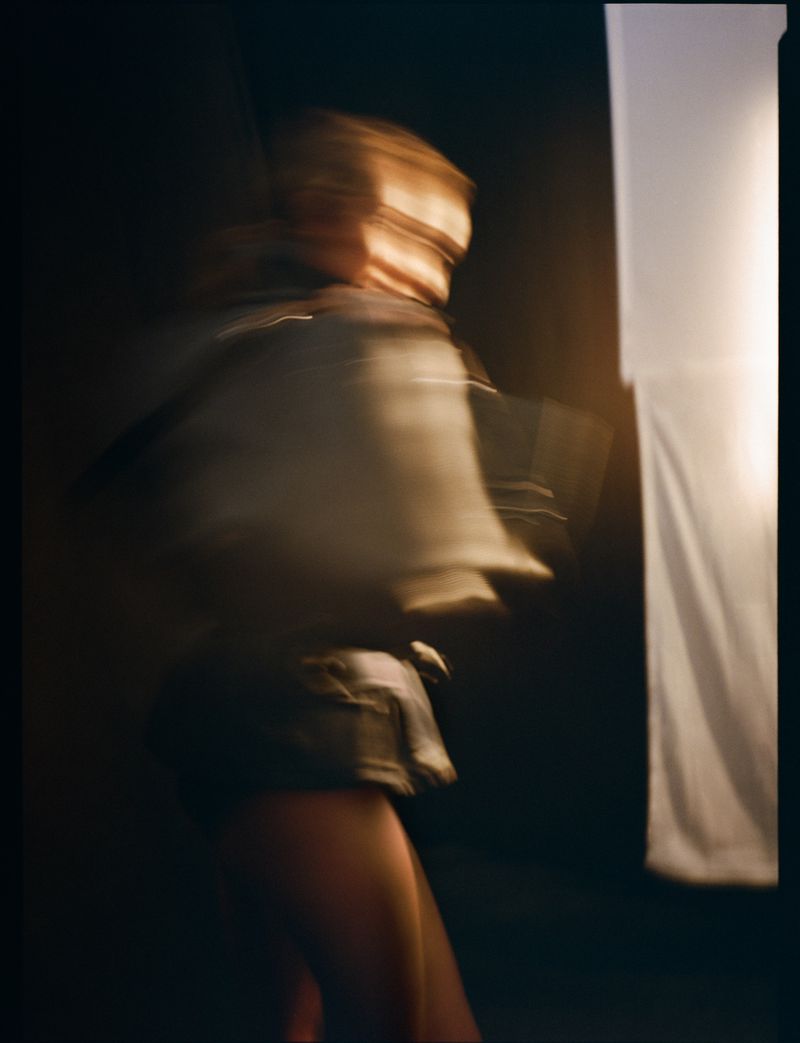 Motion blur portrait of a woman with blonde hair wearing a denim jacket and shorts, set against a dark backdrop and a brightly lit rectangle.