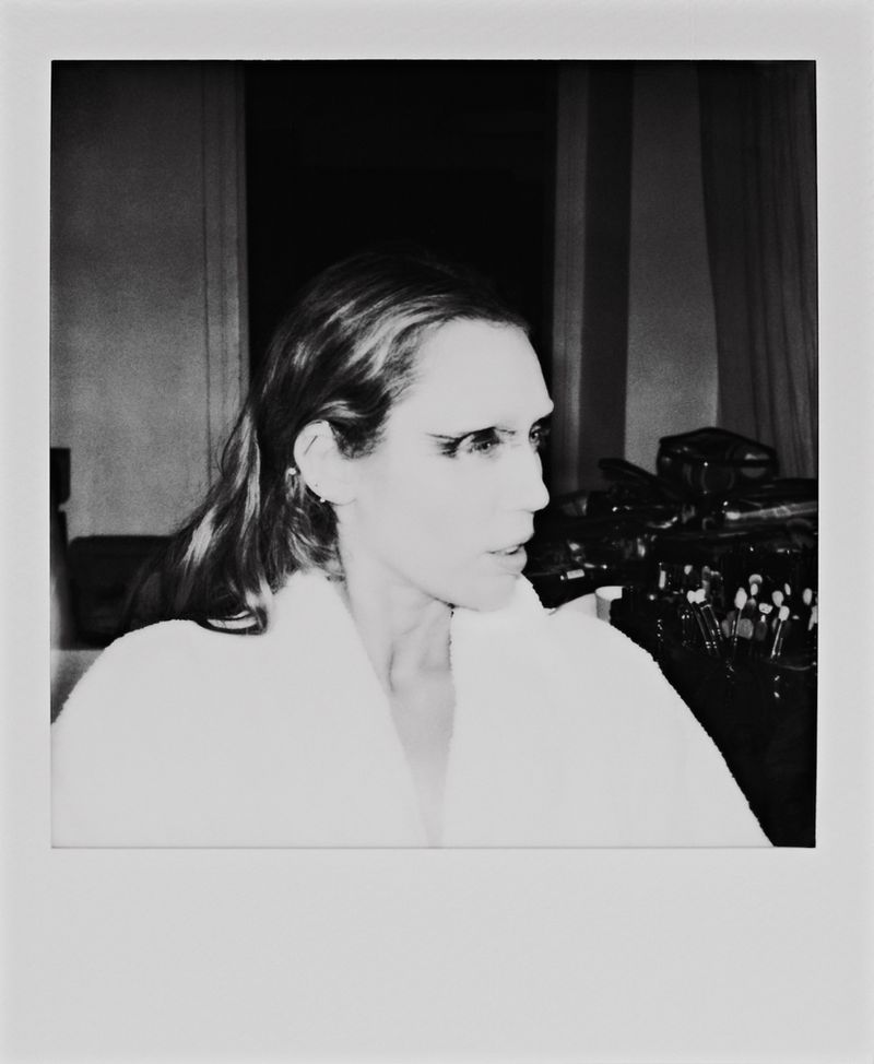Black and white portrait of a woman with dark eye makeup in a white robe, inside a room with makeup brushes and dark curtains.