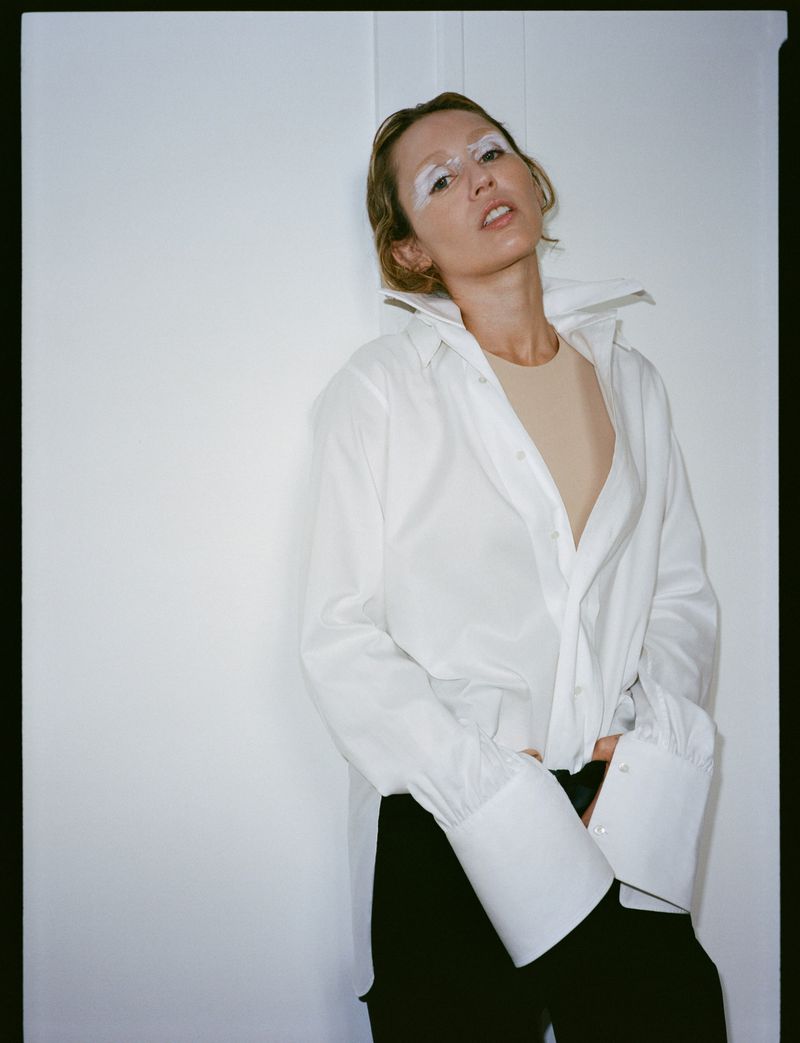 Portrait of a woman with bleached eyebrows wearing a white button-down shirt and dark pants leaning against a white wall.