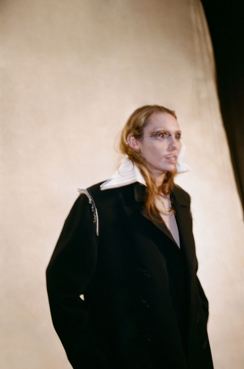 Blonde adult female model in black coat and white collar stands in a photo studio with soft, warm lighting, posing in a medium shot.