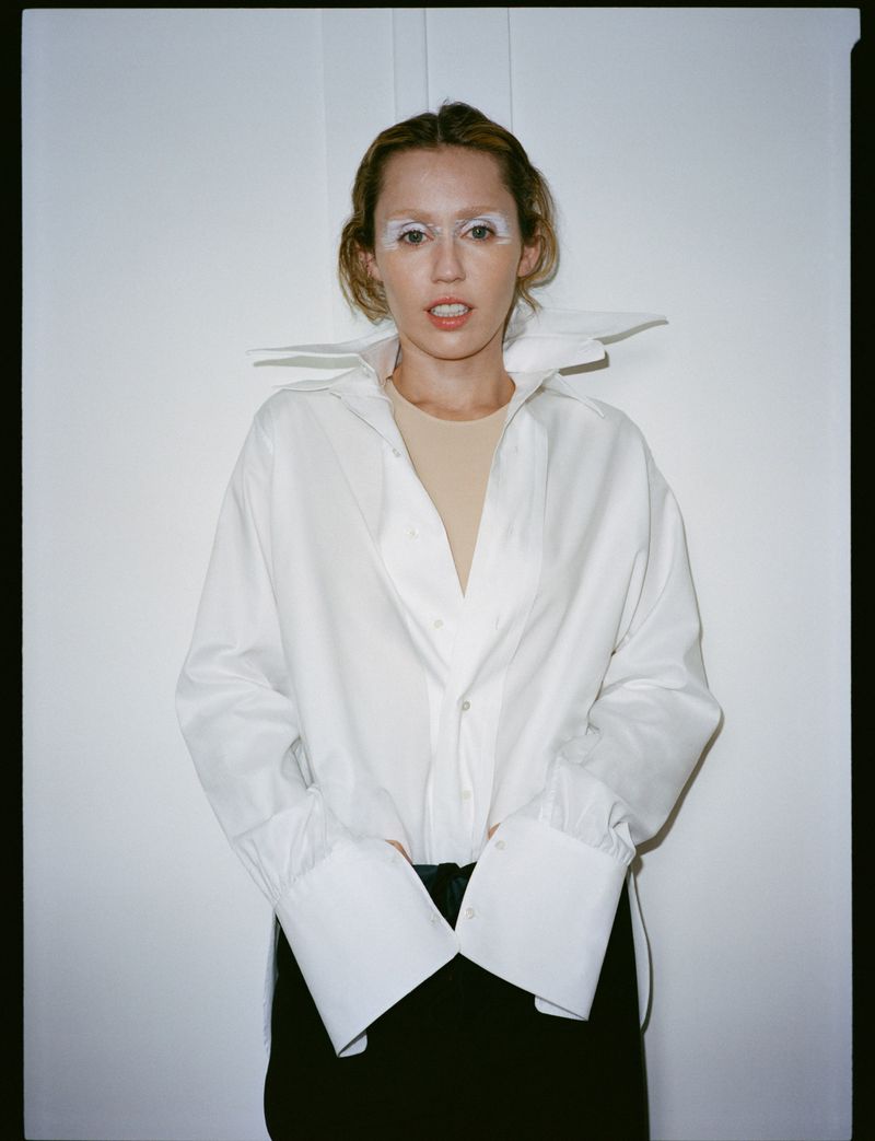 Woman in white shirt and dark skirt with white face makeup in a brightly lit studio setting, fashionable and modern.