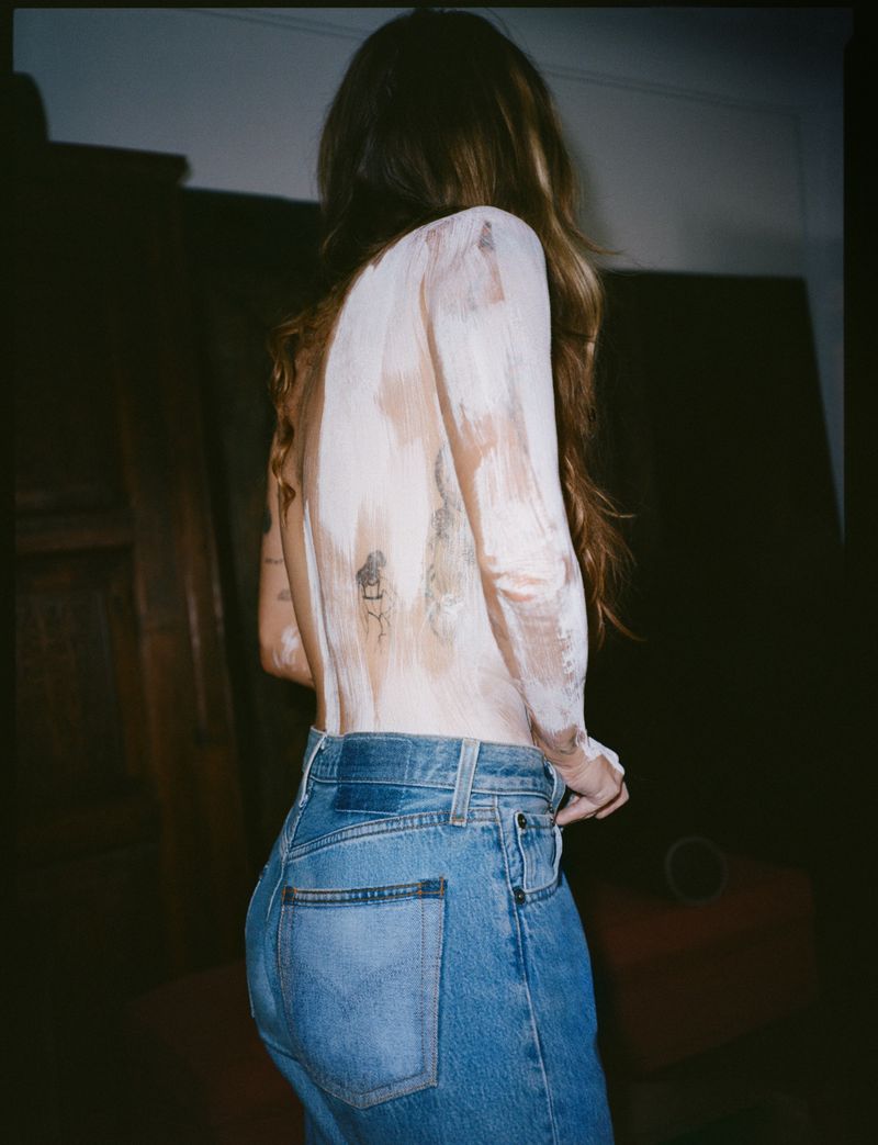 A woman with long brown hair, wearing jeans, stands indoors with her back turned to the camera. Her upper back has paint on it.
