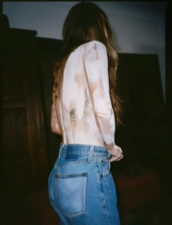 A woman with long brown hair, wearing jeans, stands indoors with her back turned to the camera. Her upper back has paint on it.