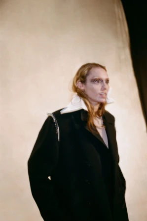Model wearing an oversized black coat with an exaggerated white collar against a warm neutral backdrop.