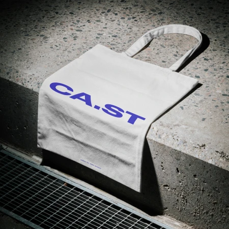 White canvas tote bag printed with bold purple CA.ST lettering draped over a rough concrete step edge.