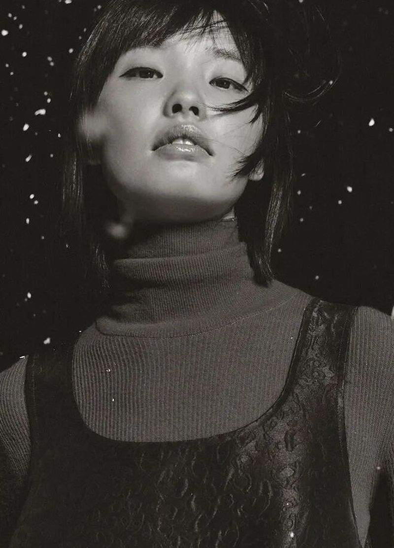 Monochrome portrait of a young woman with short hair, wearing a turtleneck sweater and patterned top, against a dark, speckled background.