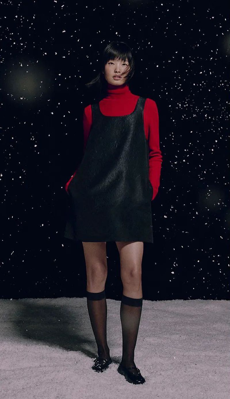 A full shot of a young Asian woman in a red turtleneck and black dress, posing in front of a black background with bokeh lights.
