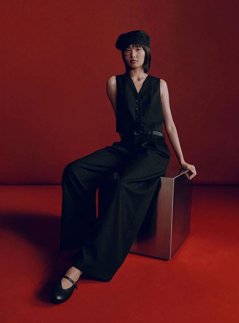 A young Asian woman in a black vest and wide-leg pants sits on a silver cube against a red backdrop, radiating understated style.