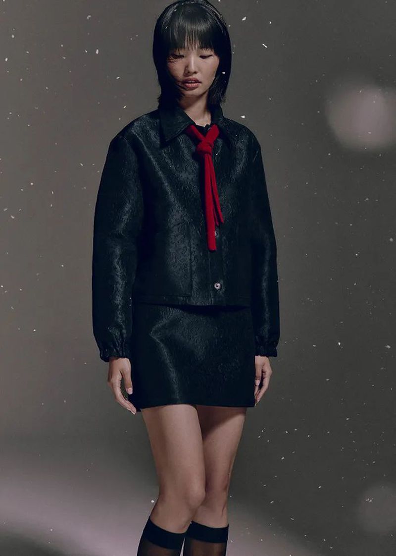 Studio shot of an Asian female model wearing a black jacket and skirt with a red scarf, posing against a gray background.
