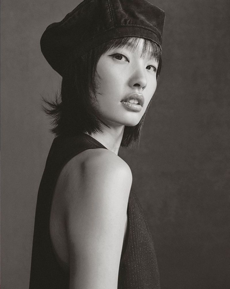 Monochrome portrait of a young Asian woman with dark hair, wearing a black beret and vest against a gray background in soft lighting.