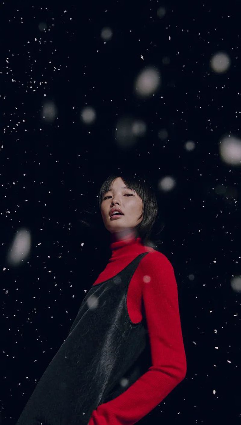 Stylish woman in red turtleneck and black dress in a studio environment with a black background and simulated snow falling.