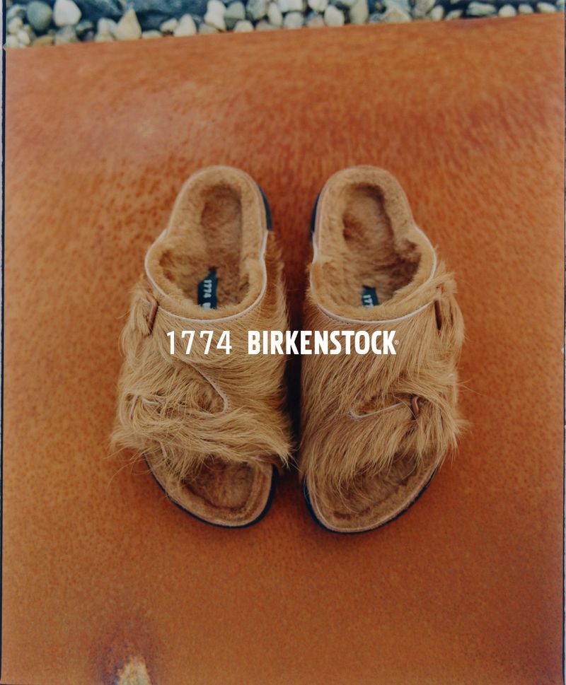 Two furry brown Birkenstock sandals are displayed on a smooth, tan leather surface, with small rocks visible in the background.