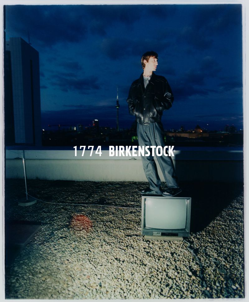 Adult male stands on a TV on a rooftop at night with city lights. He is wearing a black jacket and grey pants in an outdoor shot.