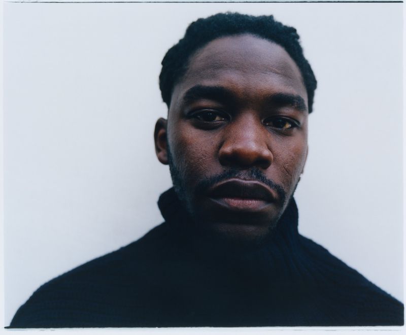 Close-up portrait of a Black adult male with short dark hair and a mustache, wearing a black turtleneck against a light grey background.