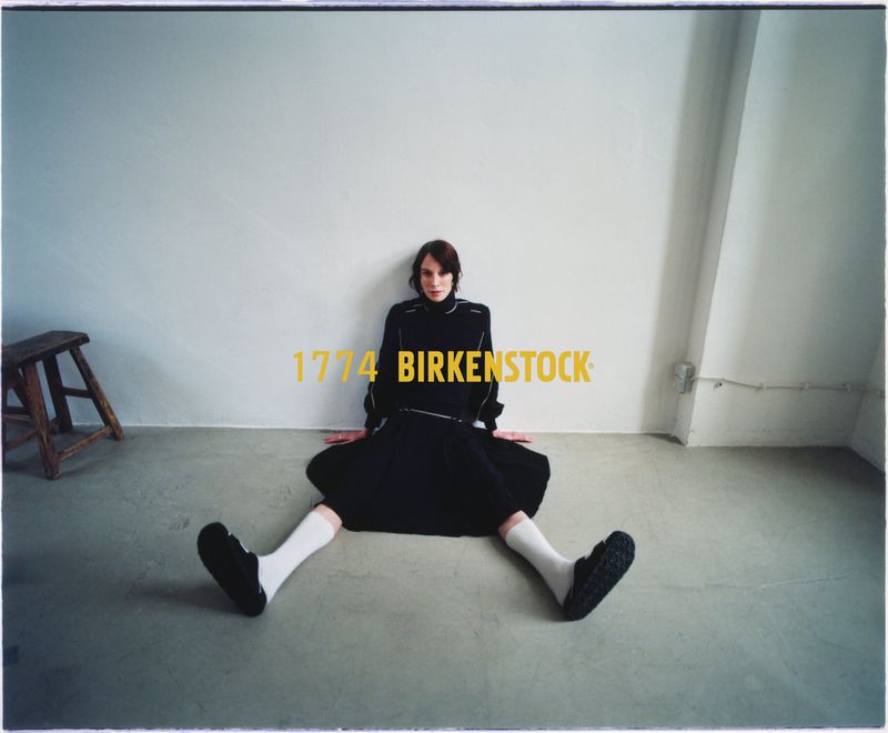 Indoor portrait of a woman sitting on the floor wearing a black dress, socks, and shoes next to a wooden stool and "BIRKENSTOCK" text.